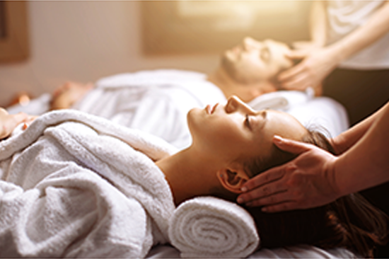 Couples massage, hands massaging each person's head as they lay peacefully on the massage table, both persons wearing a terry cloth robe.