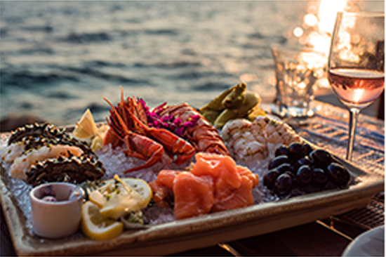 Seafood on a tray with container for sauce, lemon slices, fresh raw samon, block olives, all on ice, glass of blush wine and glass of water, sitting on a wooden table top, over looking calm waterers.
